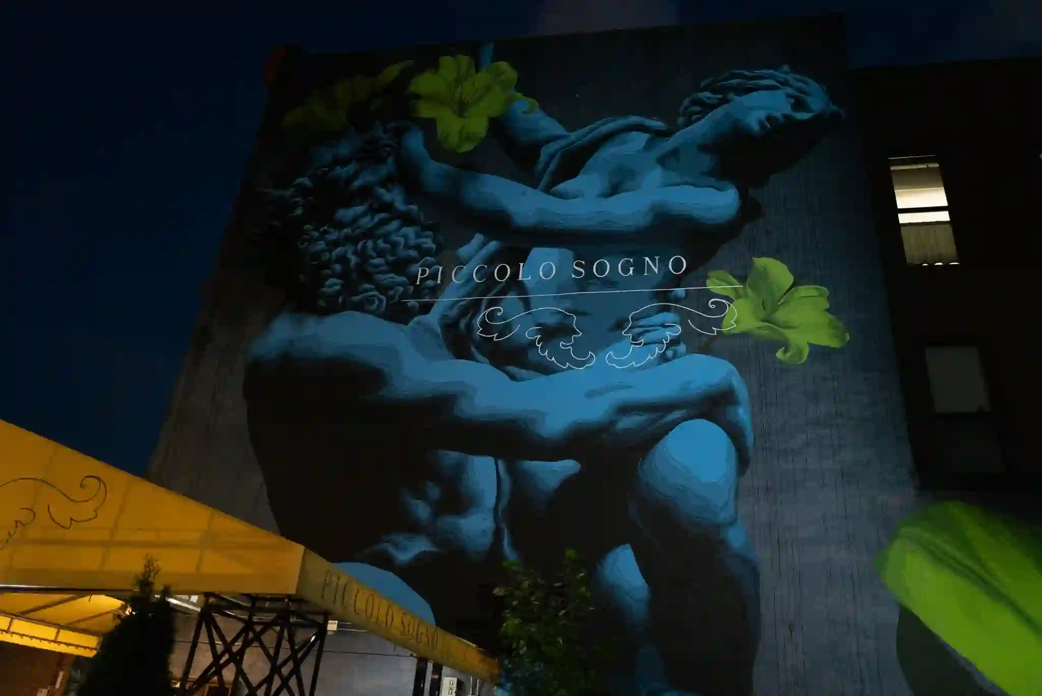 The Piccolo Sogno mural lit blue at night above the yellow awning